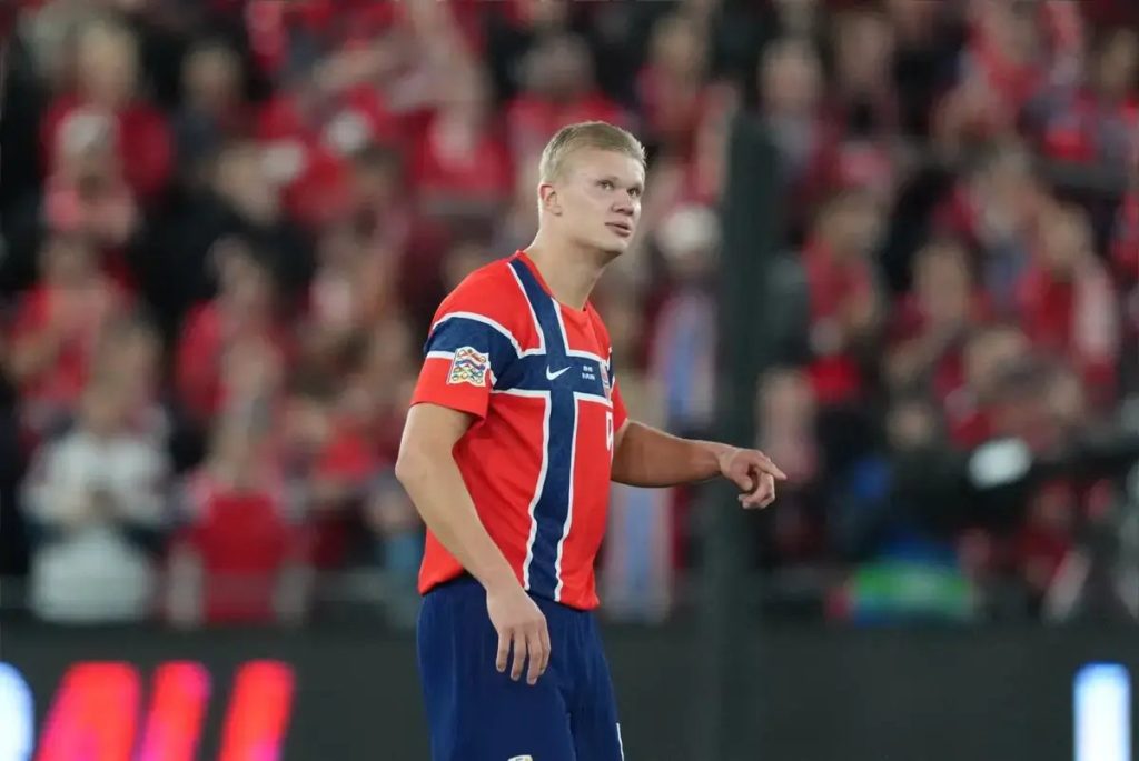 Footballer in red and navy Norway kit with white trim, looking up on a pitch as red-clad fans fill the stands behind him