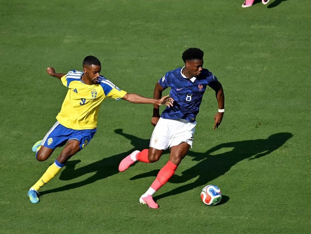 Player in blue France kit (number 6) dribbles past yellow Sweden defender (number 3) on a sunny pitch; ball at feet.