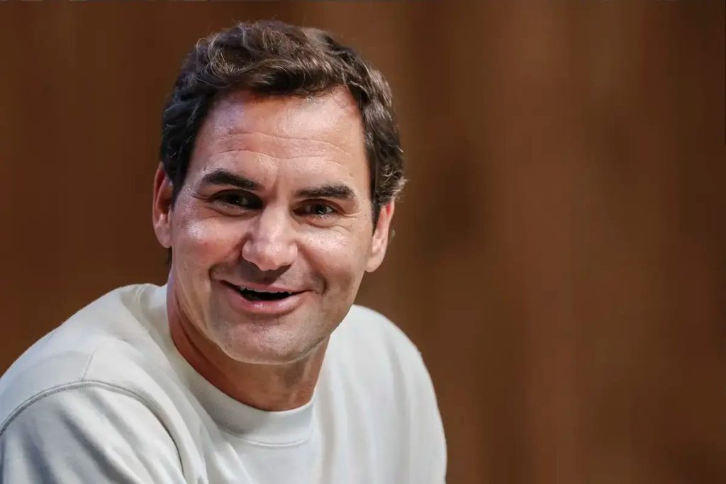 Smiling man with short brown hair in a cream sweatshirt against a warm brown wooden background, seemingly at a press or fan event.