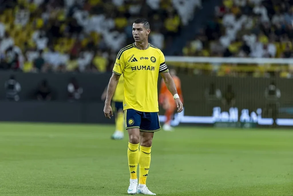 Jogador de futebol de amarelo e azul num estádio, com a braçadeira de capitão e o equipamento do Al Nassr, durante um jogo.