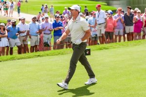 A golfer in a white cap, grey polo and white shoes walks on a green fairway holding a driver, with spectators watching behind a rope.