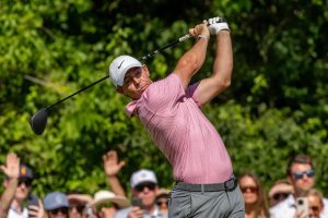 Golfer in pink patterned polo and white cap swings a driver during a tournament, with spectators watching behind him
