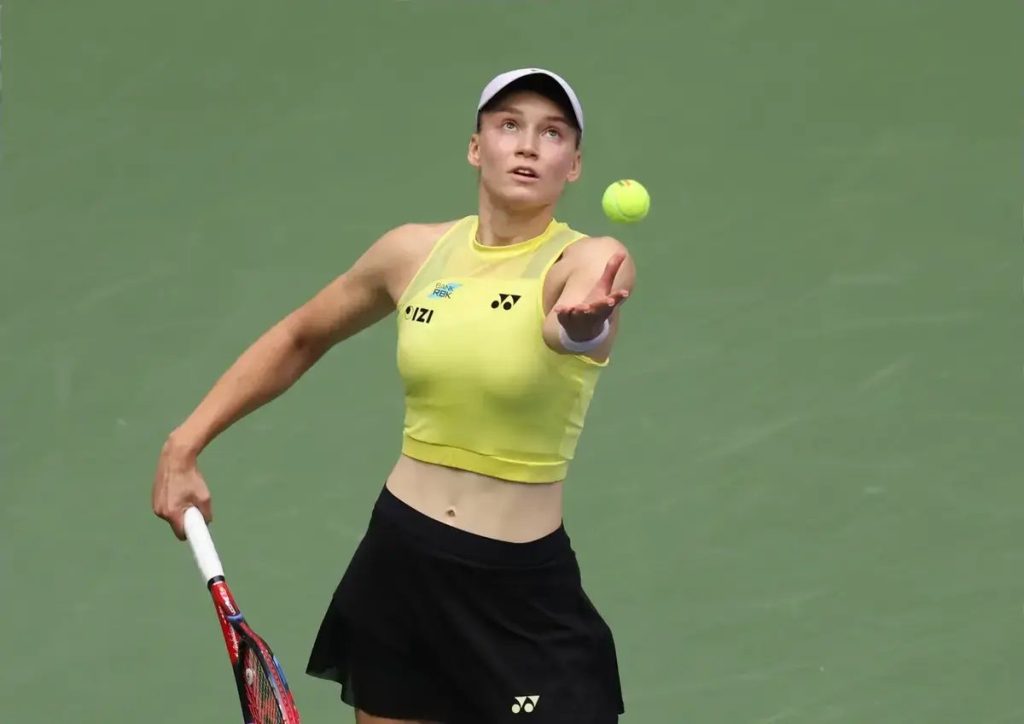 A tennis player in a yellow Yonex top and black skirt tosses a ball overhead to serve on a green hard court.