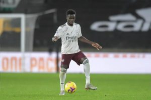 Bukayo Saka in Arsenal's white away kit dribbles a ball on the pitch during a match.