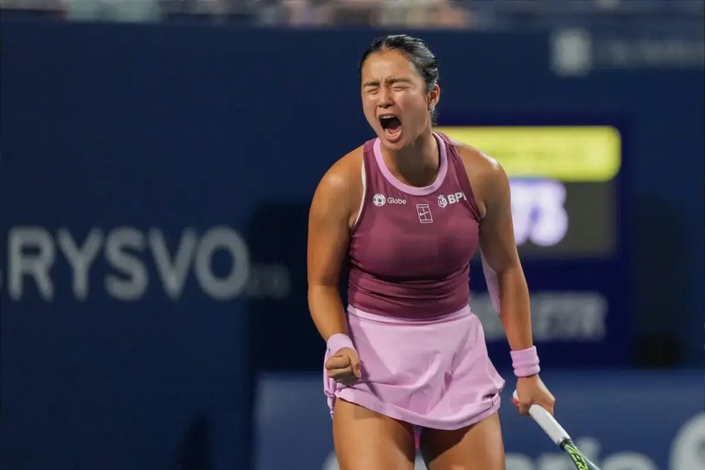 Alexandra Eala celebrating with a fist pump and yelling during a tennis match in a pink skirt and maroon top at a night event.