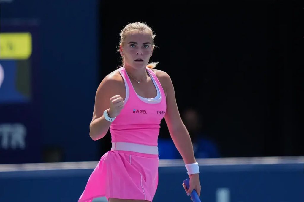 Tennis player in pink dress clenches fist in celebration on court, with a blue backdrop and a visible "AGEL" sponsor logo