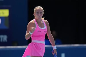 Tennis player in pink dress clenches fist in celebration on court, with a blue backdrop and a visible "AGEL" sponsor logo