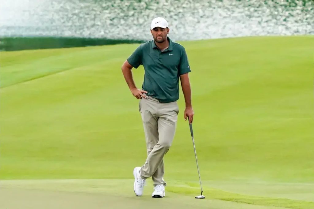 A golfer in a dark green Nike polo, white cap and khaki trousers stands with a putter beside a green, with water behind him at the 2026 Presidents Cup.