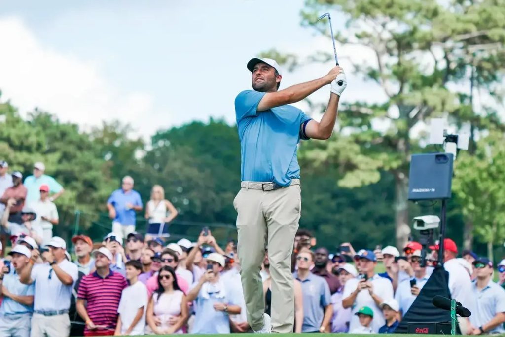 Golfer in a light blue polo and khaki pants takes a tee shot as spectators watch on a tree-lined course