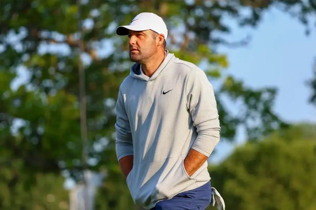 Man in a white cap and gray Nike hoodie with hands in pocket, looking off to the side on a golf course with trees behind him