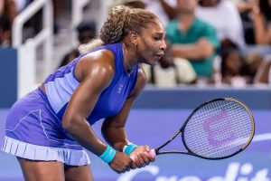 Tennis player in purple dress and blue visor-style top, braided ponytail, holding a racket mid-rally at an outdoor hard court event