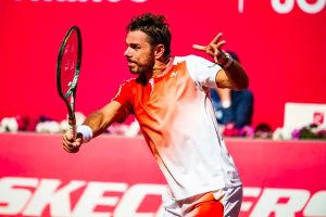 Tennis player in white and orange Yonex shirt swings a forehand on a red court, Skechers signage behind him.