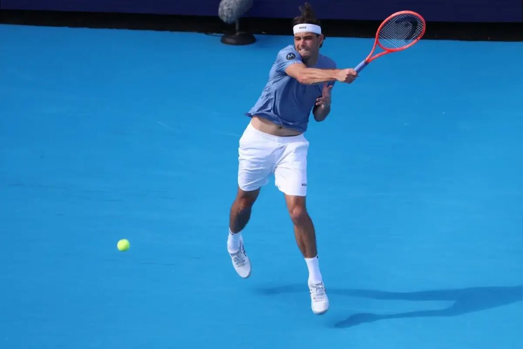 Tennis player in blue shirt and white shorts hitting a forehand on a blue court, with a red and black racket and a yellow ball in flight