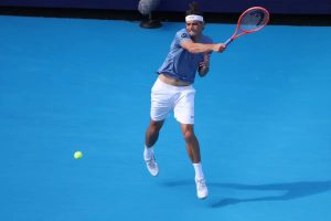 Tennis player in blue shirt and white shorts hitting a forehand on a blue court, with a red and black racket and a yellow ball in flight