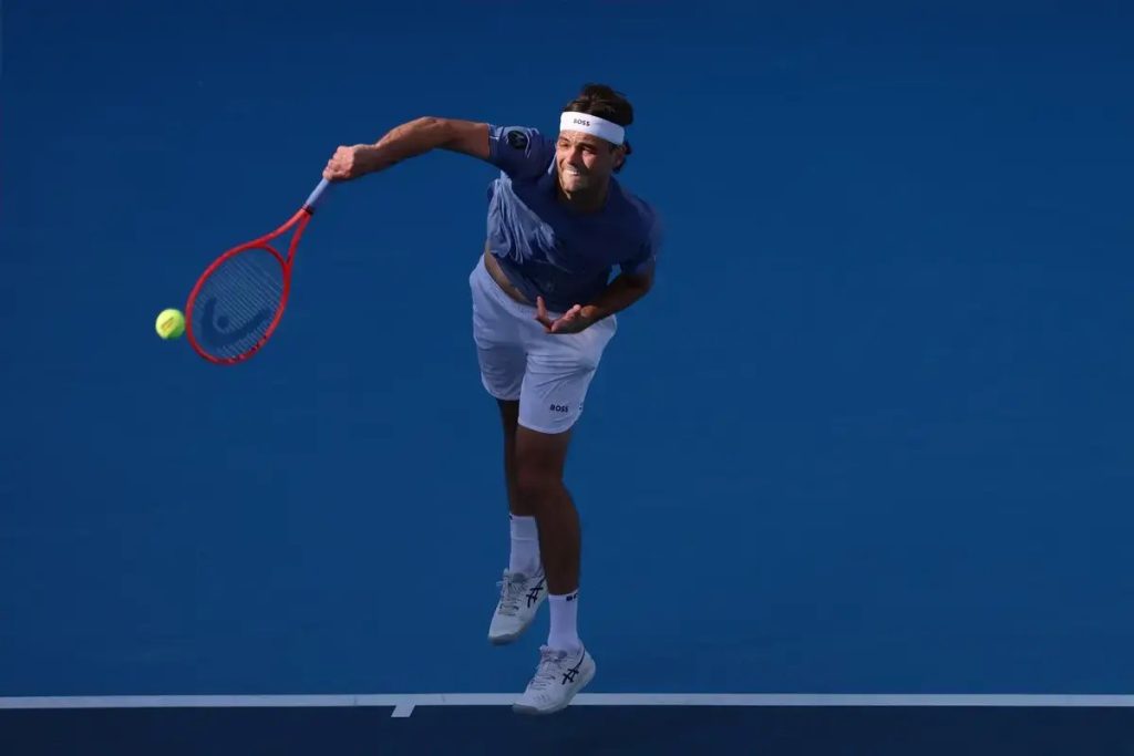A tennis player in a blue shirt and white shorts serves with a red racquet on a blue court, ball in the air.