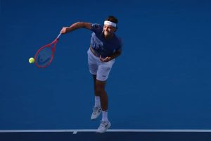 A tennis player in a blue shirt and white shorts serves with a red racquet on a blue court, ball in the air.