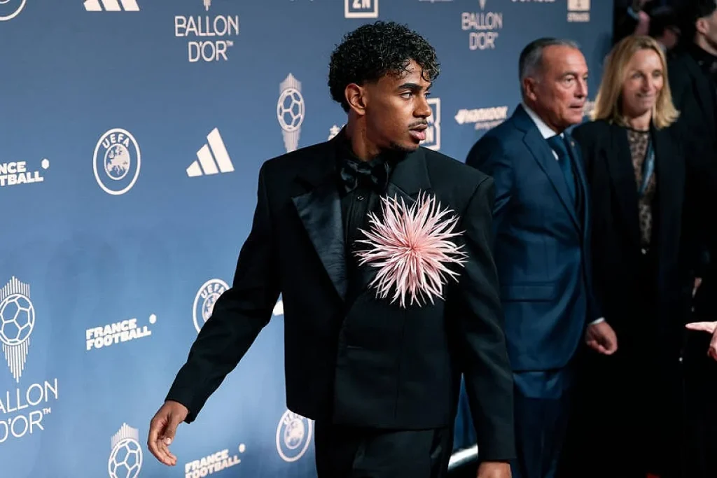 Jovem de cabelo encaracolado, fato preto e flor cor-de-rosa ao peito, na gala do Ballon d'Or, com fundo azul da UEFA e France Football.