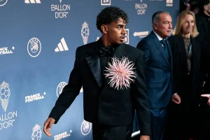 Jovem de cabelo encaracolado, fato preto e flor cor-de-rosa ao peito, na gala do Ballon d'Or, com fundo azul da UEFA e France Football.
