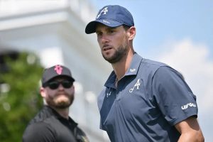 Golfer in navy cap and LIV Golf polo at a tournament, with a security staffer in sunglasses behind him