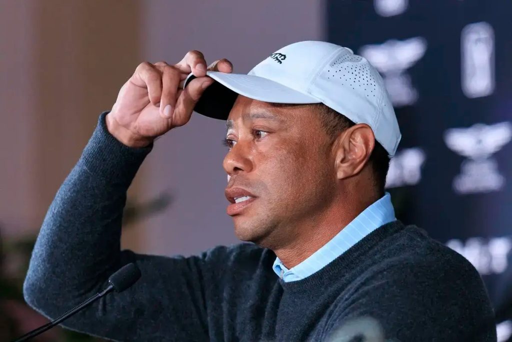 A golfer in a white cap with a grey sweater adjusts his hat at a press conference, with a dark backdrop bearing golf tournament logos.