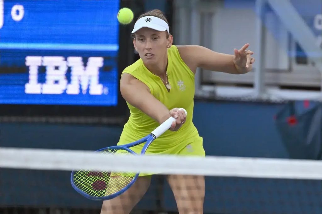 Tennis player in a yellow sleeveless outfit and white visor hits a forehand return during a match, with an IBM sponsor board behind her.