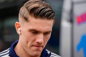 A young man with slicked-back hair and white earbud in one ear, wearing a navy and white Arsenal-style training top, looks downward beside a white vehicle