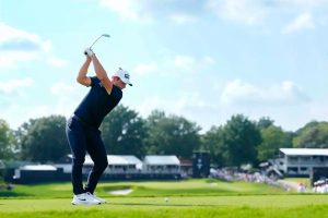 A golfer in a navy shirt and white cap swings a club on a fairway, with a crowd and clubhouse behind him