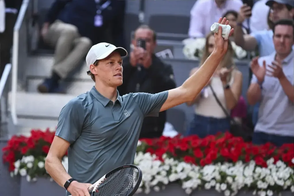 Tennis player in a grey polo and white cap raising his hand and holding a racket, acknowledging fans at a clay-court event with red and white flowers.