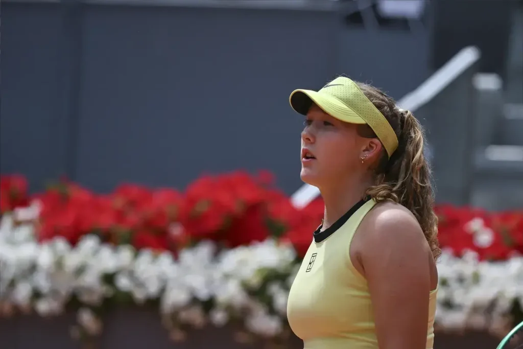 A tennis player in a yellow visor and yellow top with black trim looks up during a match, red and white flowers behind her on a clay court.