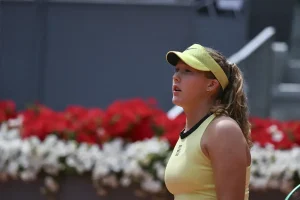 A tennis player in a yellow visor and yellow top with black trim looks up during a match, red and white flowers behind her on a clay court.