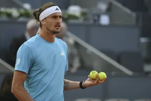 Tennis player in light blue Adidas shirt and white headband holding two yellow balls on a clay court, Madrid setting