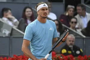 Tennis player in a light blue shirt and white Adidas headband holding a racket on a clay court, with spectators and red flowers behind him
