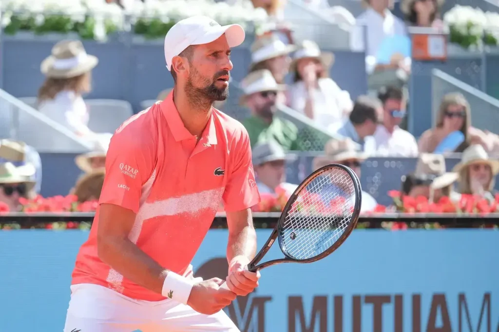 Tennis player in white cap and coral Lacoste polo with Qatar Airways sponsor, gripping racket on clay court at Mutua Madrid Open.