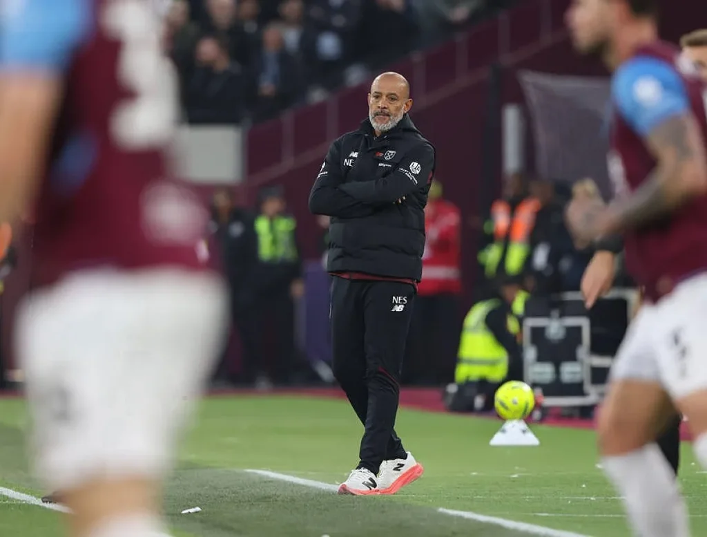 Treinador de fato preto com as mãos cruzadas à beira do relvado, em jogo do West Ham; jogadores desfocados em primeiro plano.