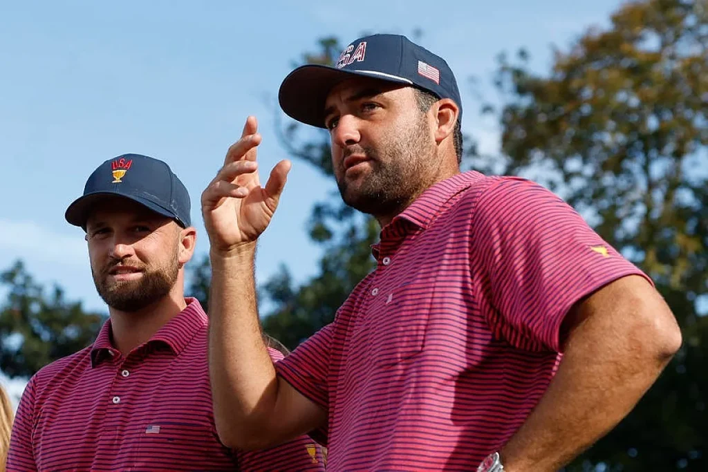 Dois golfistas com bonés azul-marinho com "USA" e vestem polos rosa às riscas; o da frente gesticula, em campo de golfe.