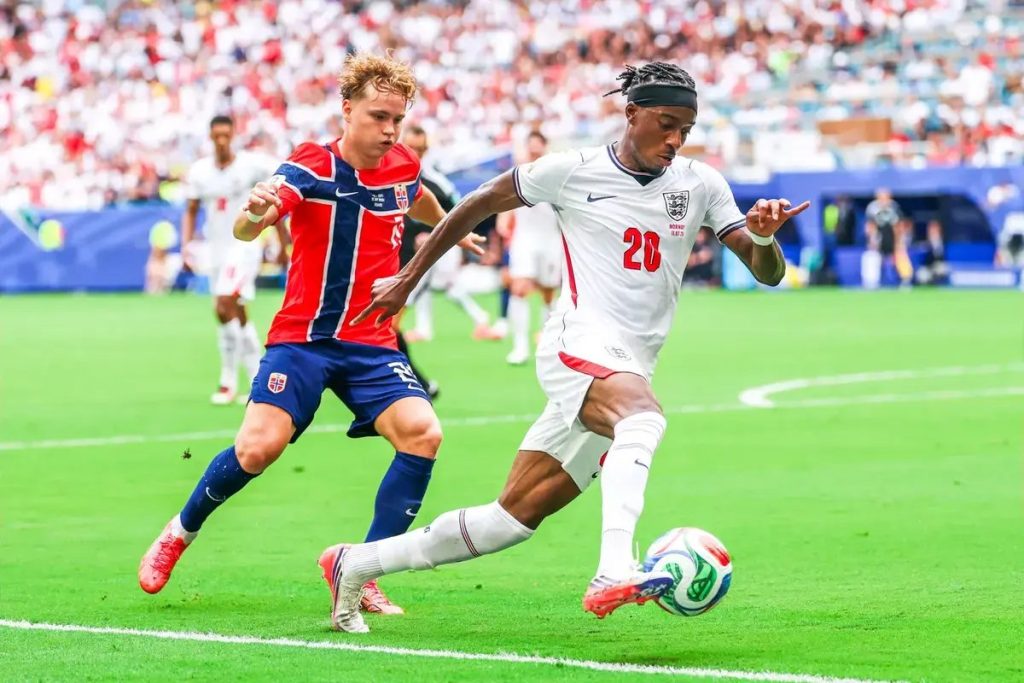 Player wearing white England No. 20 kit dribbles past a Norway defender in red and blue during a match, the article's subject Noni Madueke.