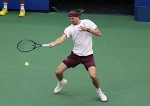 Tennis player in a white shirt and maroon shorts hitting a forehand on a green court at the US Open, with a yellow ball in flight.