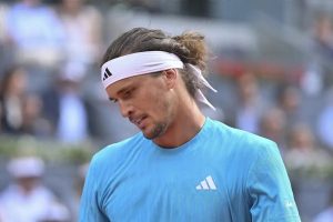 Tennis player in a light blue Adidas shirt and white headband, head bowed on a clay court with blurred spectators behind him.